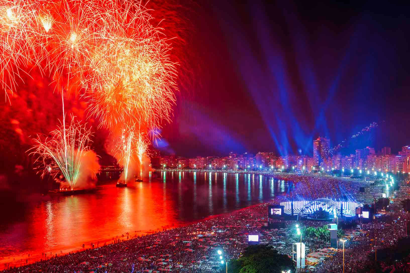 Red and gold fireworks exploding over a crowded beach at night with illuminated cityscape and searchlights in background